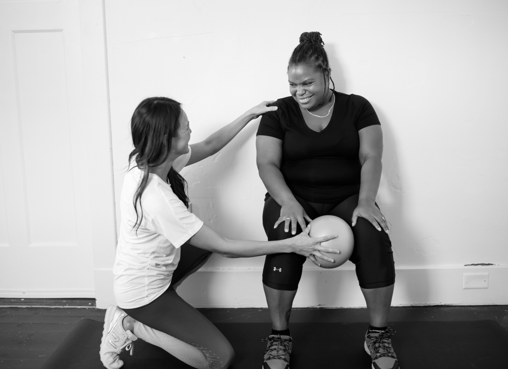 Patient leans against the wall in a wall sit with a ball in between her knees. PT kneels next to her