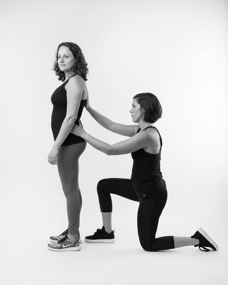 One person kneels behind a standing person with her hands on the standing persons hips
