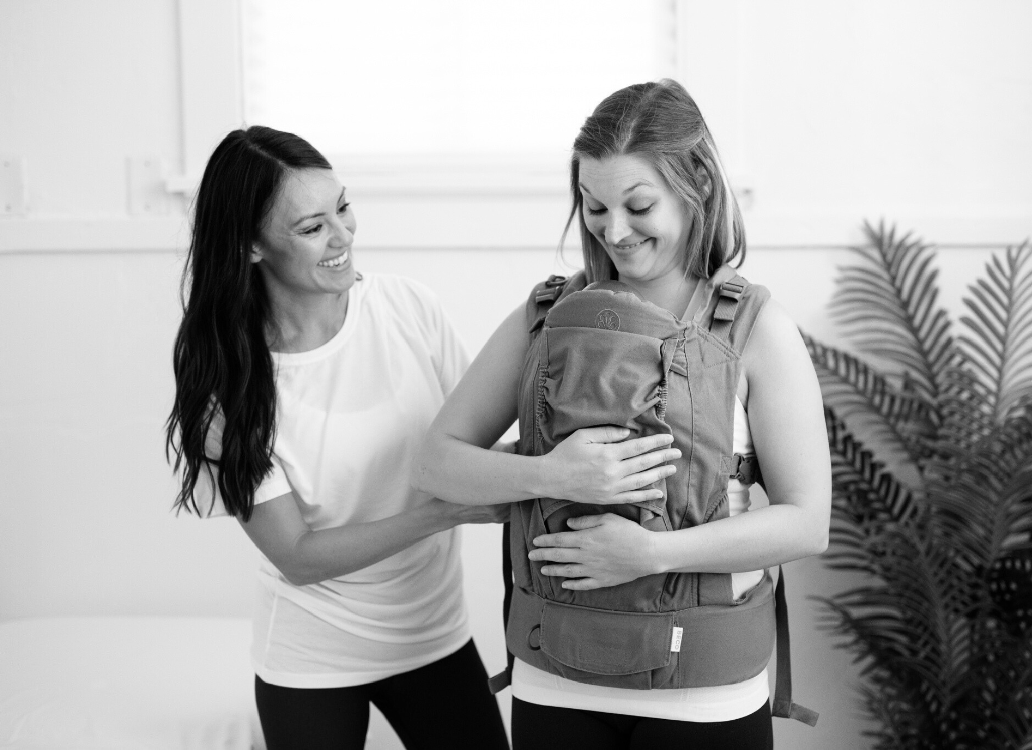 mom holds baby in a carrier on her chest, physical therapist stands next to her with her hand on her back