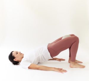 woman is holding bridge pose with a grey pilates ball between her thighs.