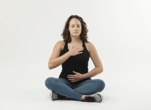 Woman sits on ground cross legged with one hand on heart, one hand on belly, eyes closed. 