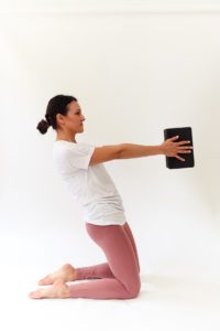 woman is holding yoga block between hands while slightly tilted back in an upright position on her knees.