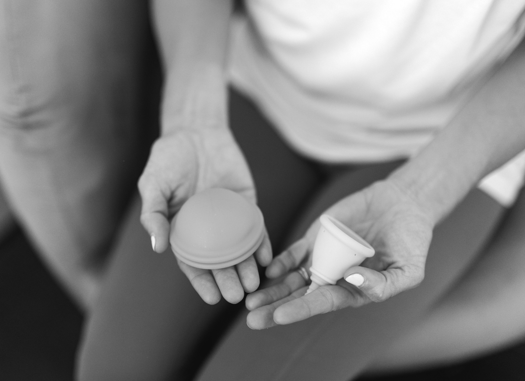 close up of woman sitting in chair holding a menstrual cup in one hand and a menstrual disc in another