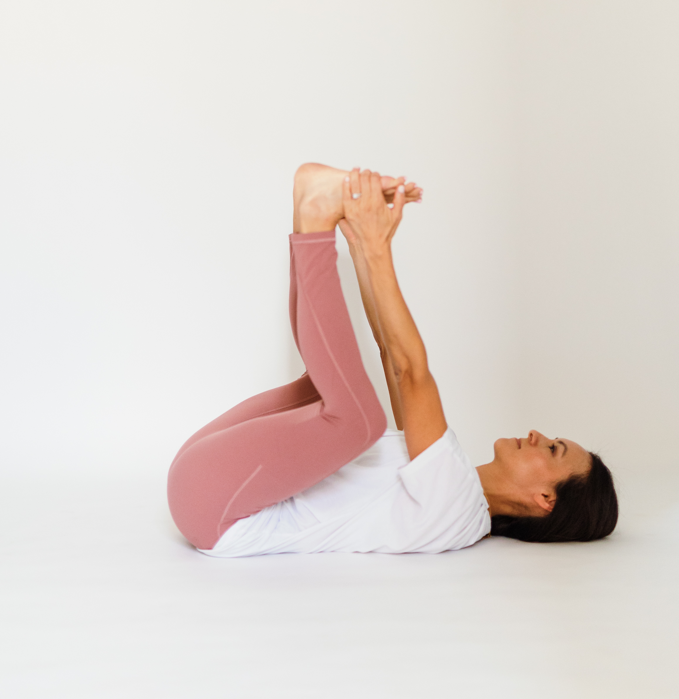 Woman lies on ground on her back with legs in the air, holding onto legs in happy baby pose.