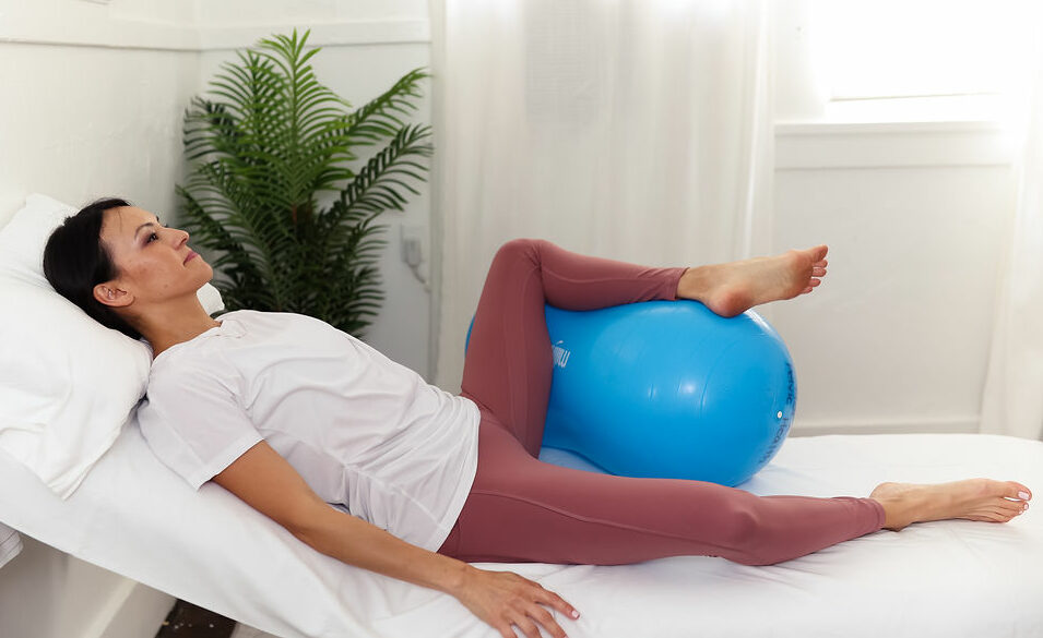 Woman lays on exam table with one leg propped up under exercise ball